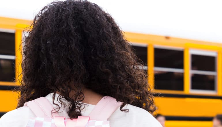A teen girl walks to a yellow school bus while wearing a backpack.