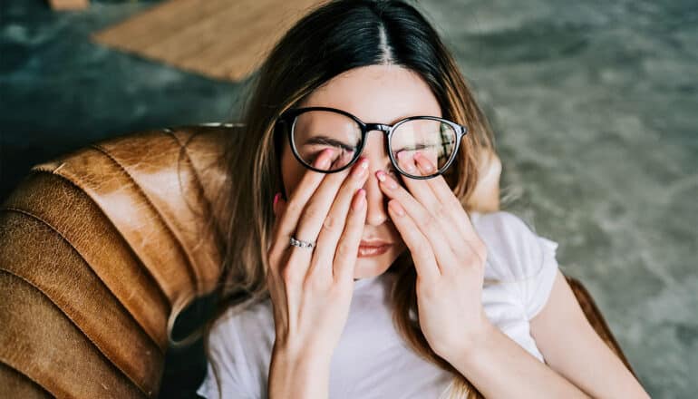 A young woman in a chair rubs her eyes under her glasses.