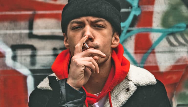 man in red hoodie and coat squints while smoking cigarette