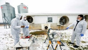 three people in white coveralls stand outside barn with equipment