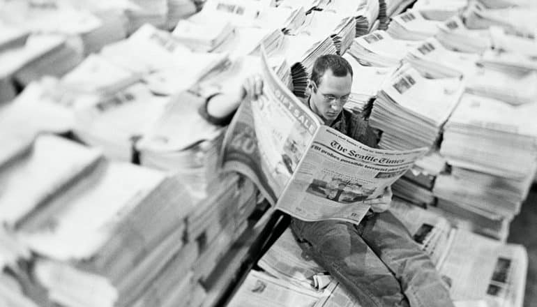man reads newspaper amid stacks - journalism