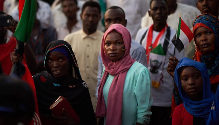 protestors in Sudan - woman in pink scarf and green top stands in crowd