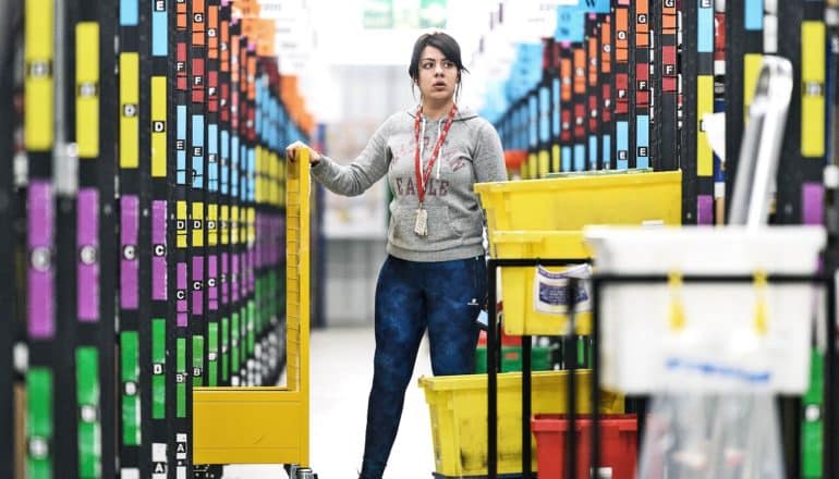 An Amazon warehouse worker looks slightly confused as she stands in between rows of organized and color coded products while holding a rolling bin