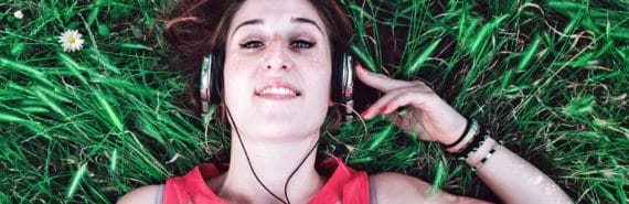 woman lies in grass with one arm raised to hold silver headphones on her head - listening to podcasts