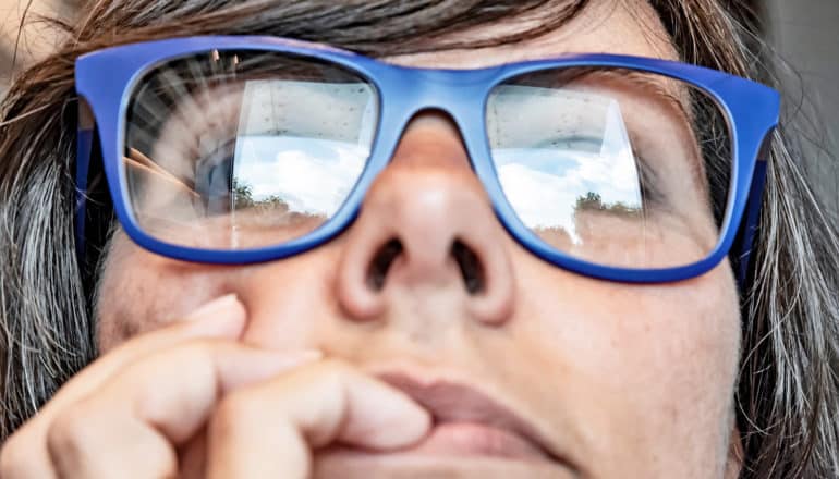close-up of person's face, hand to mouth pensively, blue eyeglasses with reflection of white square