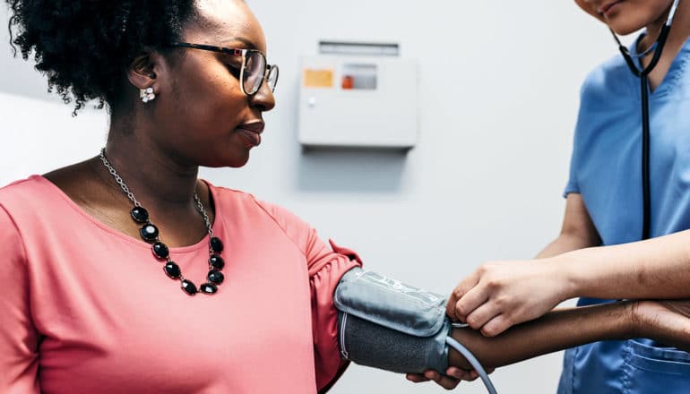 A pregnant woman in orange is at the doctor getting her blood pressure taken by a nurse in blue scrubs