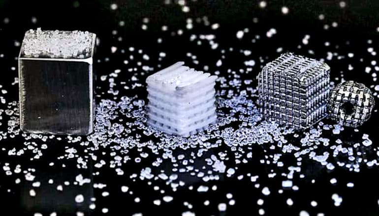 A block of metallic-looking magnesium covered in salt sits next to one of the 3D-printed salt templates, a small, white block with visible pores, and one of the bone implants that looks like the salt template, but is made of shiny magnesium metal. Around the 3 blocks, there are grains of salt on a black background.