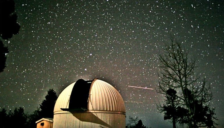 The domed building housing the telescope and the silhouettes of several large trees frame a view of the star-filled night sky.