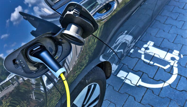 A navy blue electric car has the gas cover open with a line recharging the car and the symbol for electric car charging on the ground next to the car