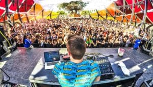 A DJ in a blue shirt faces away from the camera and towards a large crowd of dancing concert-goers between two red and orange barriers