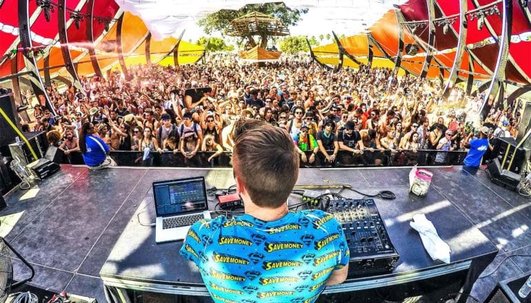 A DJ in a blue shirt faces away from the camera and towards a large crowd of dancing concert-goers between two red and orange barriers