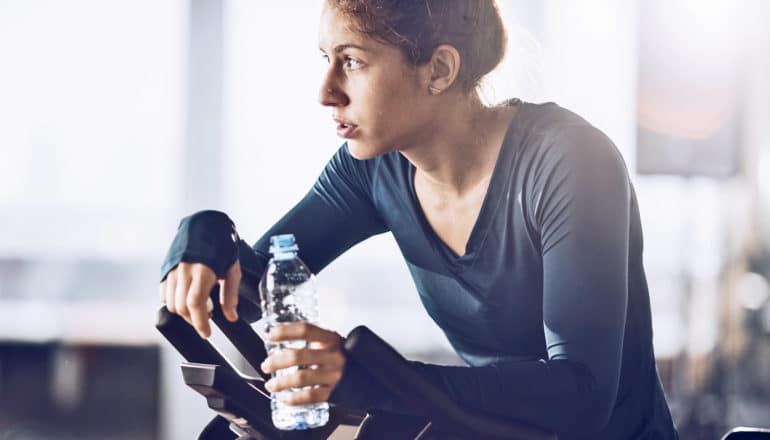 sweaty woman with neutral expression rests on stationary bike holding bottle of water