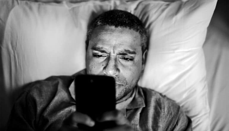 The image shows a man in bed looking at his phone with frustration.