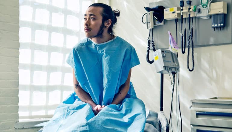 worried-looking genderqueer person sits on exam table in blue gown