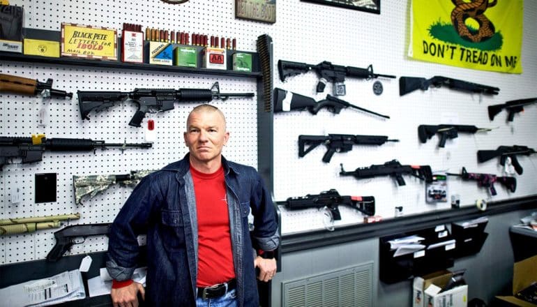 The gun shop manager, wearing a red t-shirt with a blue over-shirt, leans back on a counter in front of a white wall displaying a range of guns