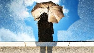 A person holds an umbrella while looking down at their reflection in water, which shows the sky behind them is clear and blue