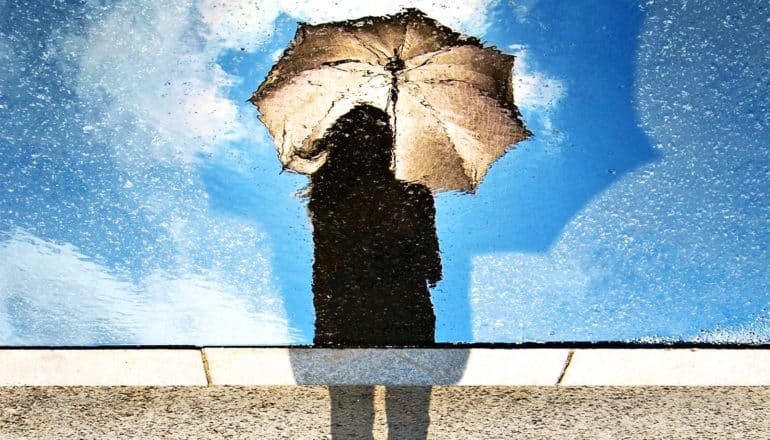 A person holds an umbrella while looking down at their reflection in water, which shows the sky behind them is clear and blue