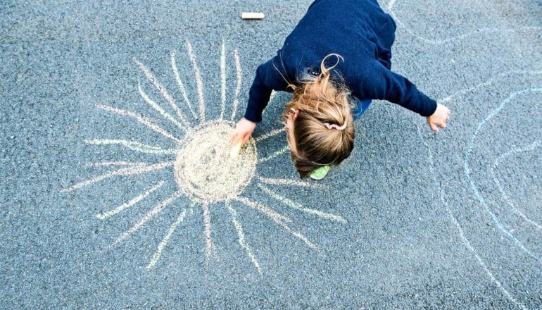 A young kid in a navy shirt uses chalk to draw a yellow sun on some asphalt