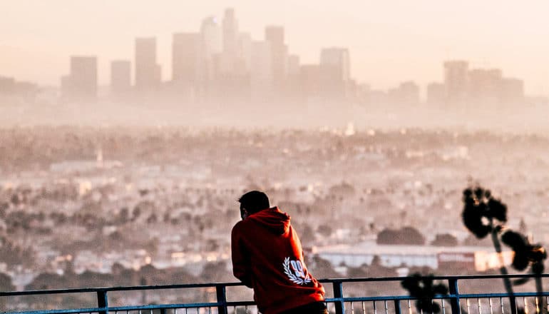 A man in a red hooded sweatshirt looks over a hazy cityscape, with buildings obscured by pollution.