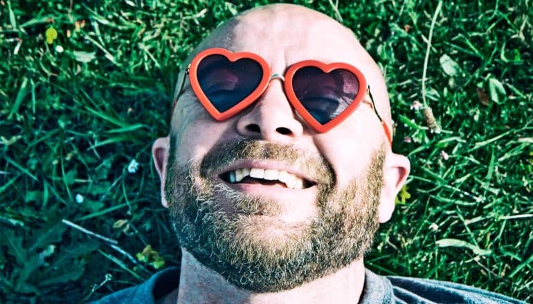 A bald man wearing heart-shaped glasses smiles with contentment as he lies on grass