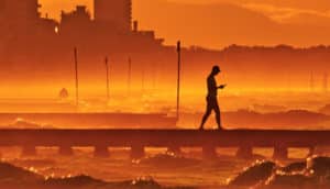 A man in profile walks looking at his phone down a pier on a hazy and hot day, a city in an orange haze behind him