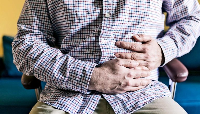 A man in a button-up shirt clutches his stomach in pain as hie sits in a chair