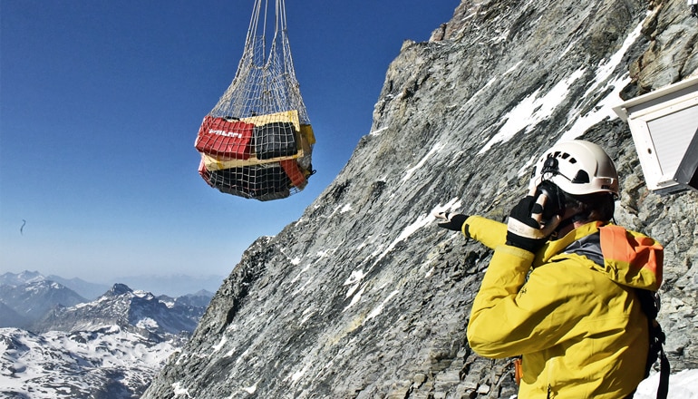 A researcher in a helmet and yellow jacket on the Matterhorn points at a suspended pack of supplies being hoisted up the mountain