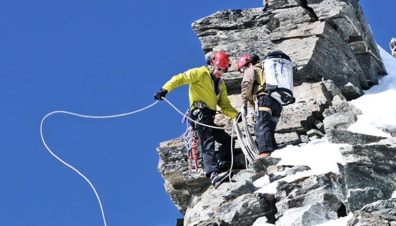 Two researchers climb the rocky face of the Matterhorn mountain wearing safety gear, with one researcher tossing a length of rope back behind them and down the mountain