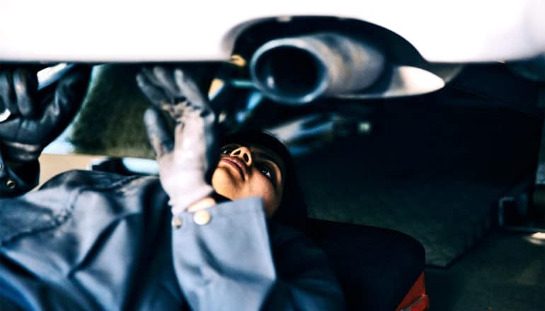 a mechanic slides under a white car lying on a rolling board near the car's exhaust system