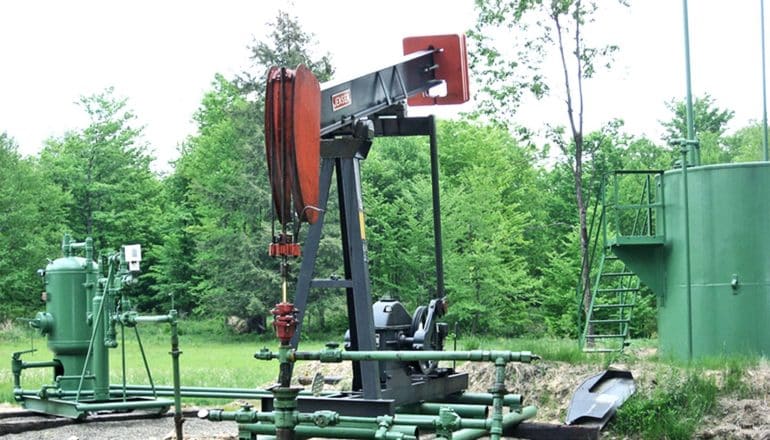 The oil and gas well is red and black and sits near green trees and painted green pipes