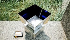 The cooling system looks like a box covered in reflective material with an open top and darkly reflective interior. The box sits on a small wall, connected to a display, in front of some grass.