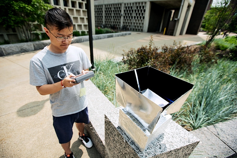 Lyu Zhou stands outside next to the boxy cooling system holding a display 