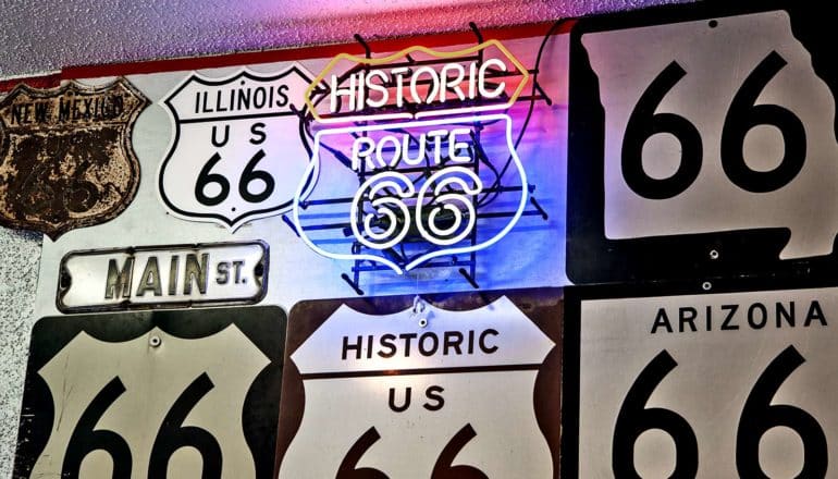 several old black and white route 66 signs hang near a bright red and blue neon version of the sign