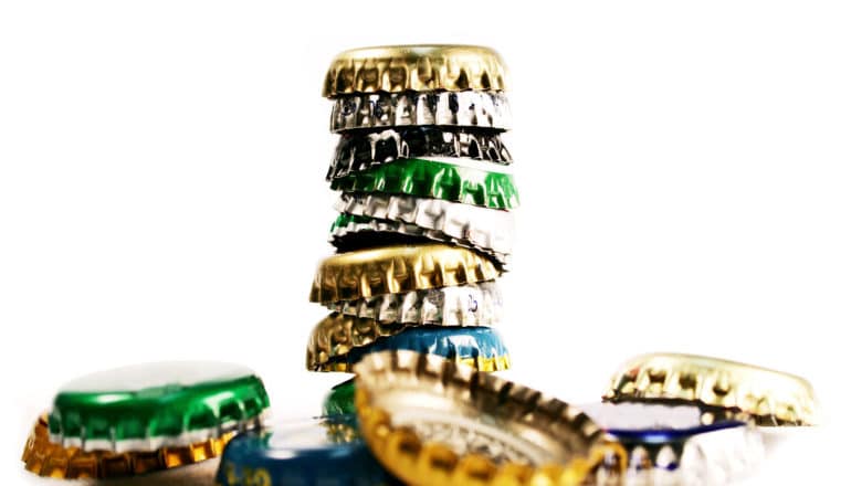 A stack beer bottle caps sits in front of some that have fallen off, all on a white background