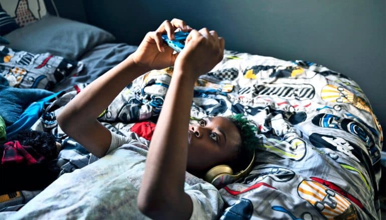 A young teen boy lies on his bed (covered in skateboarding-themed blankets) while playing a game on his phone and wearing headphones
