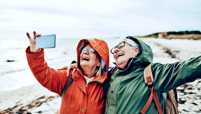 Two friends in rain coats take a selfie on the beach while laughing