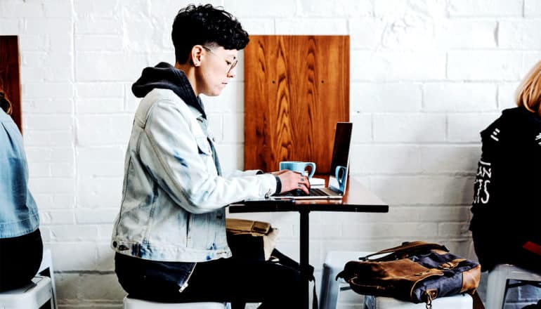 A person wearing a jean jacket and a short haircut in profile uses their laptop at a cafe table against a white wall