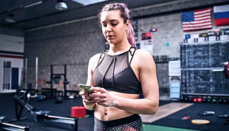 a woman in workout gear looks at her phone while at the gym, with weights and mats in the background