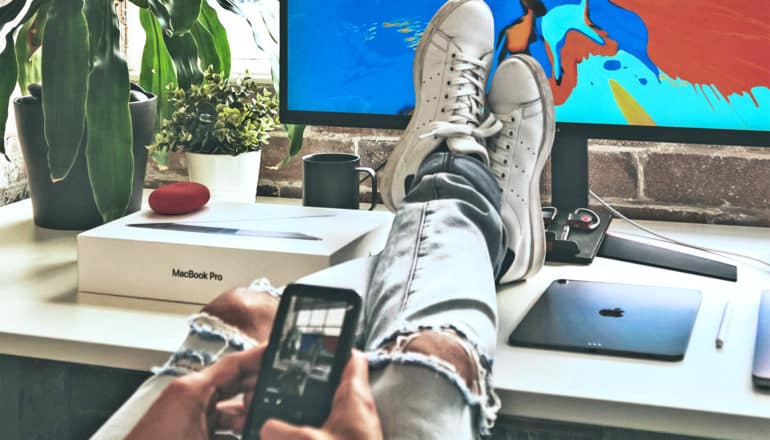 A man uses his phone while he has his feet on his desk in front of his computer monitor while wearing torn jeans and white tennis shoes.