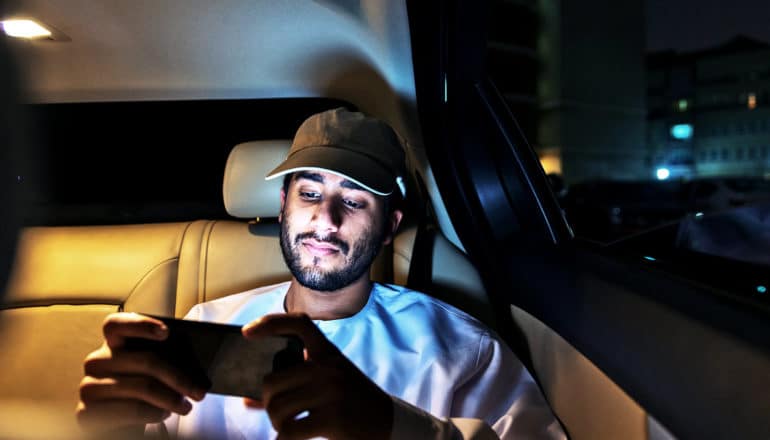 A young man wearing a baseball cap sits in the backseat of a car watching video on his phone at night.