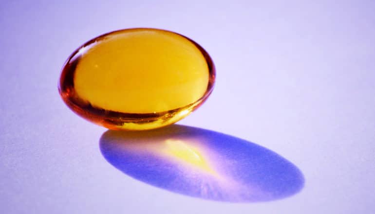 The image shows a single vitamin d pill on a purple background, with light shining through the pill. (vitamin D supplements concept)