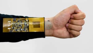 outstretched arm and fist. Golden circuitry on a rectangle forms the receiver on the arm, while a silver pattern on a small circle sits on skin of wrist