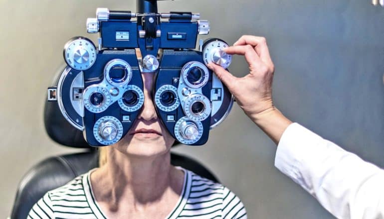A woman in a black and white striped t-shirt getting an eye test looks through the phoropter at the eye chart as the doctor reaches out to adjust the device