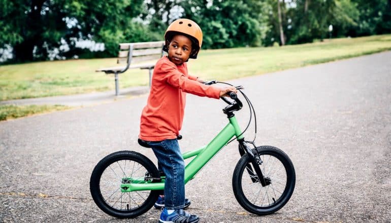 A young boy in an orange shirt and orange bike helmet looks behind him while sitting on a green bike.