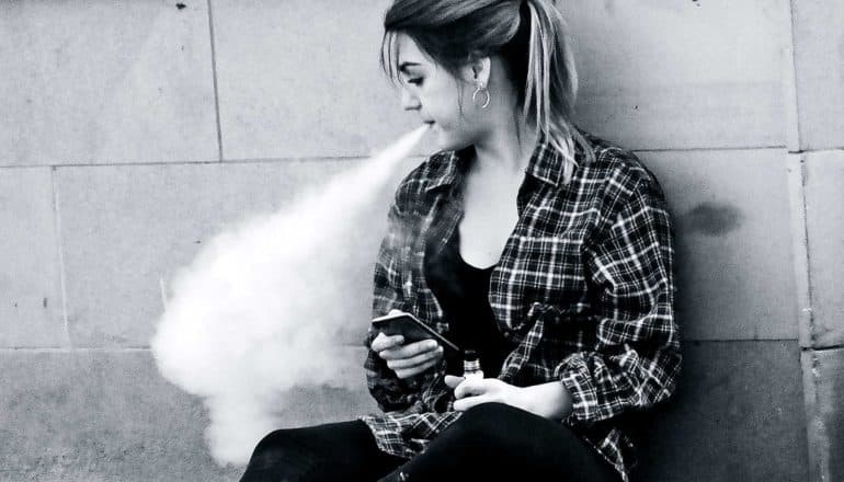 a young woman sits against a wall in a plaid shirt vaping and holding her phone