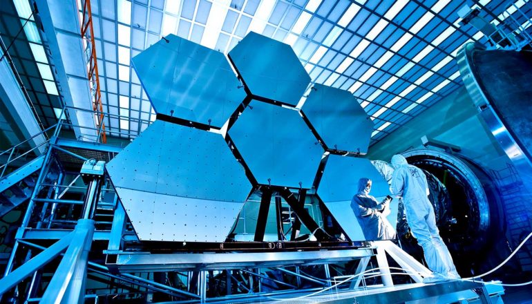 Two scientists in white clean suits work on the James Webb telescope, 6 massive hexagonal panels for which stand behind them