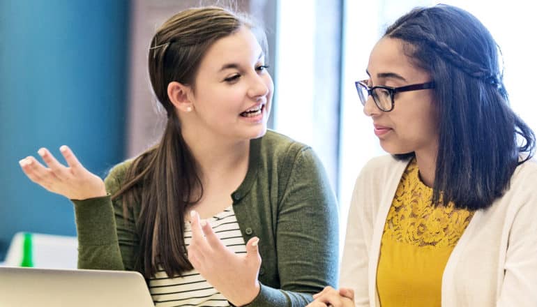 One teen in a green sweater holds up her hands while talking to a student in yellow, while they sit in a classroom in front of a computer