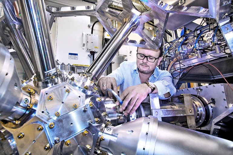 A researcher reaches towards a metallic device with a variety of tubes, wires, and pipes coming out of a central core.