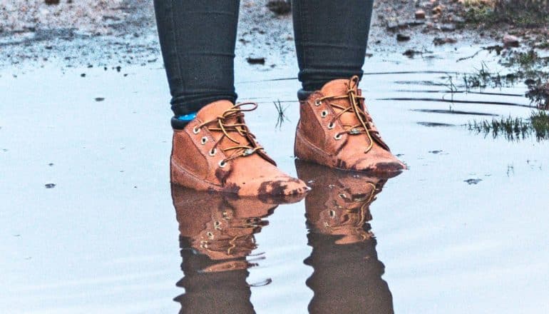 A person with boots on stands in the middle of a muddy puddle, with the water up to their ankles and some drier soil in the background