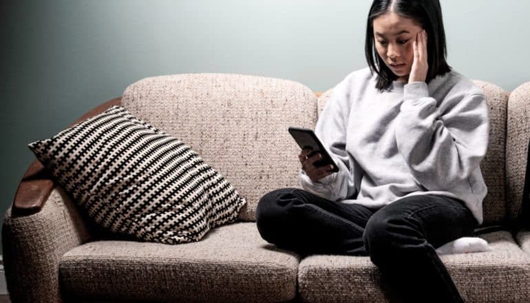 upset woman sits on couch, looks at phone with hand to forehead